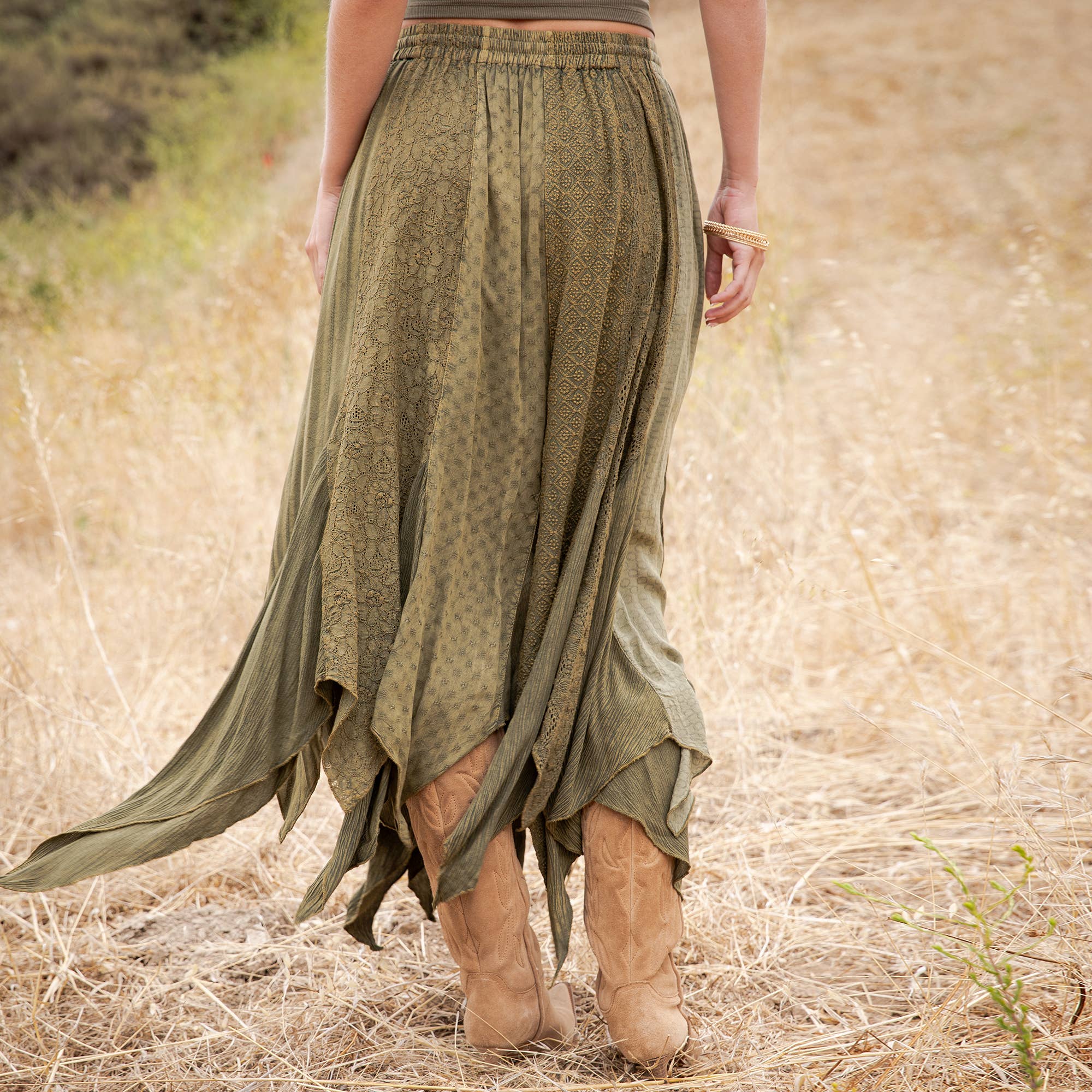 An olive green bohemian maxi skirt with a subtle tonal pattern, shown on a model from behind in a dry grass field. The skirt features an asymmetrical draped hem and is styled with tan cowboy boots.