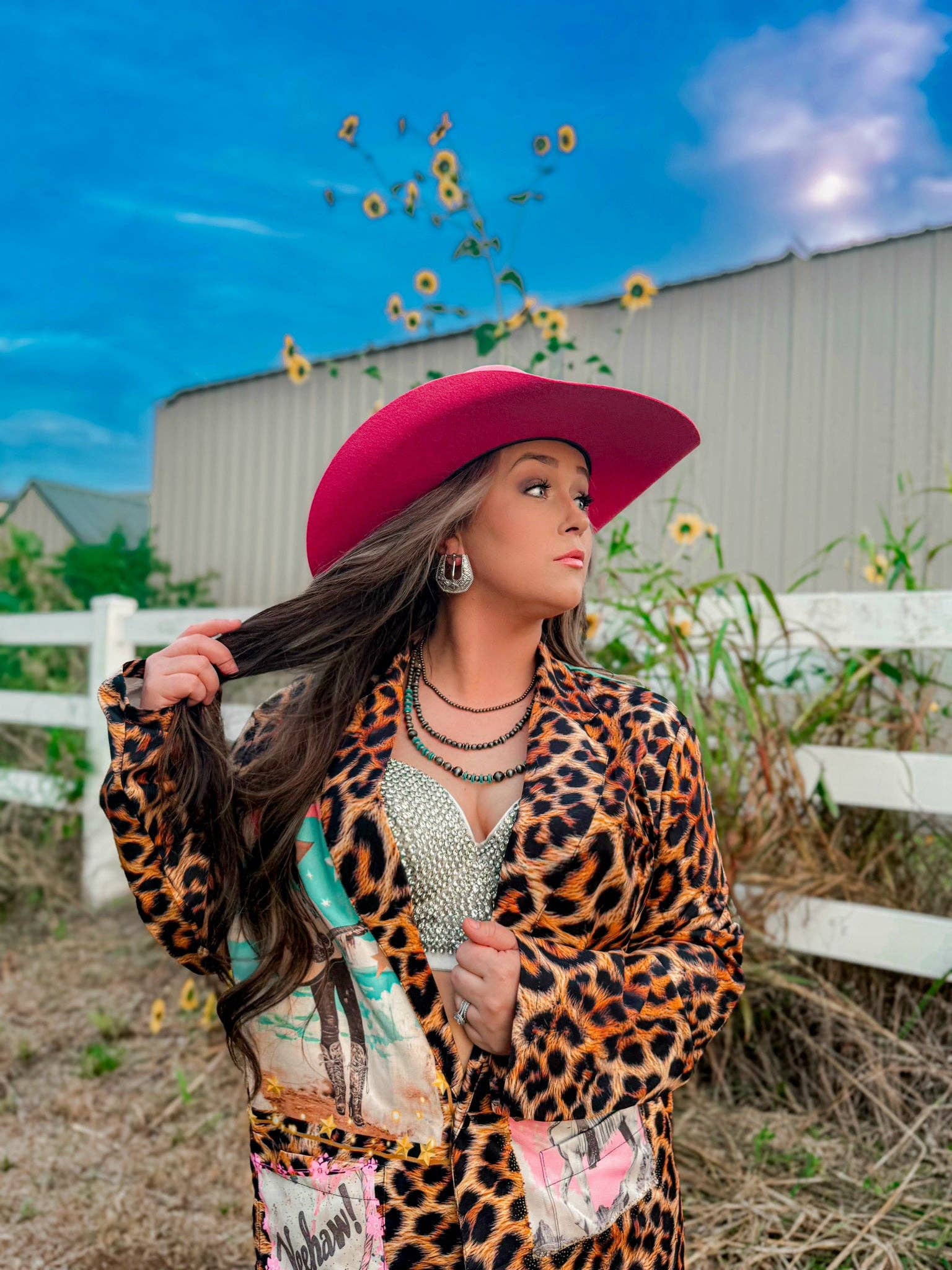 Woman in a leopard print jacket and pink hat standing outdoors with sunflowers and a white building in the background.