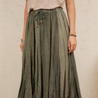 A person wearing a long, pleated skirt in a muted sage green with a subtle tonal geometric pattern. The skirt features an elastic waistband with a drawstring and small orange decorative beads.
