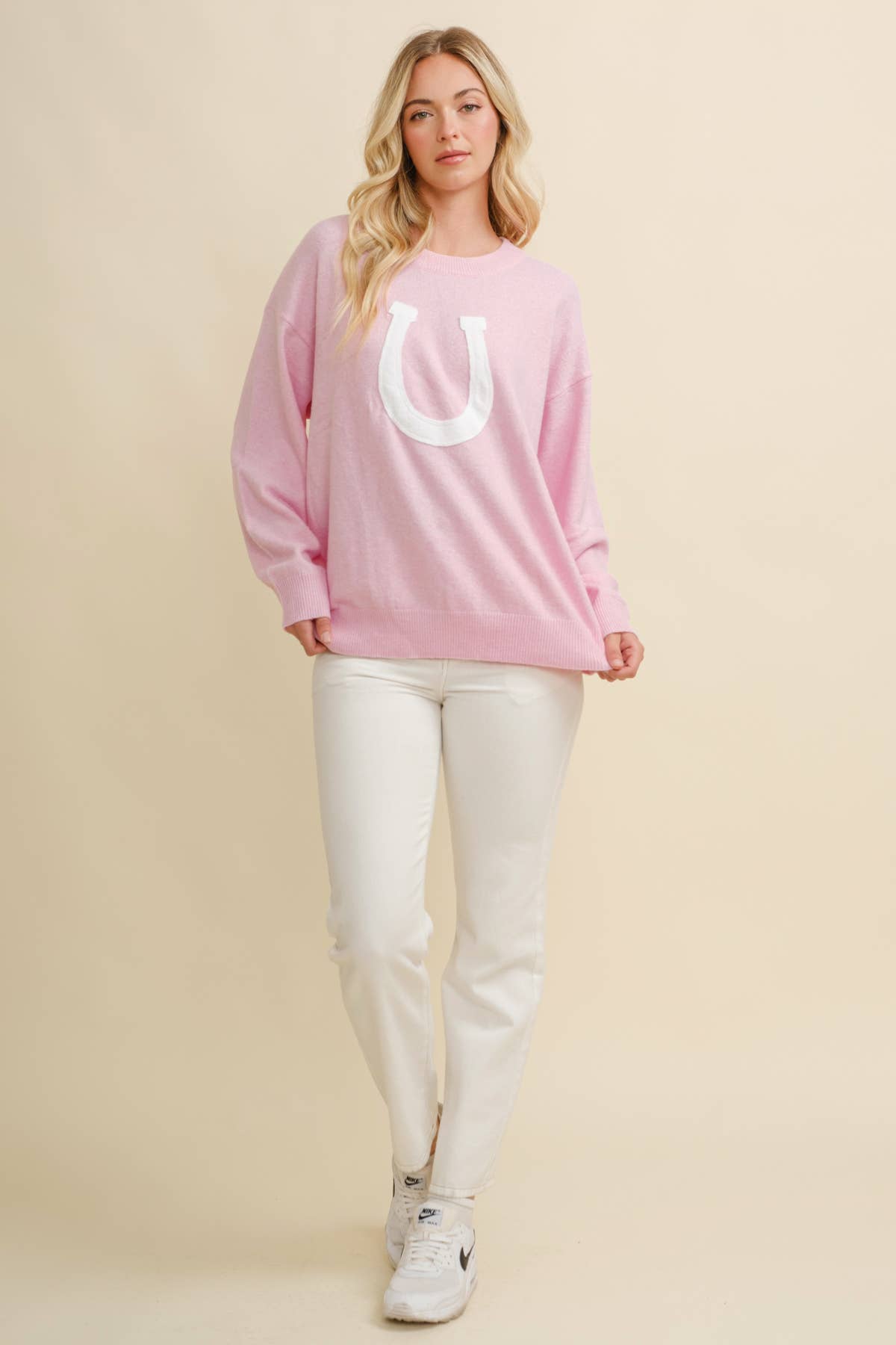 Woman wearing a pink sweatshirt with a white horseshoe and white pants on a beige background