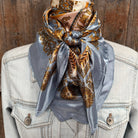 Decorative scarf with gold and brown patterns on a light blue fabric draped over a denim jacket against a wooden background.