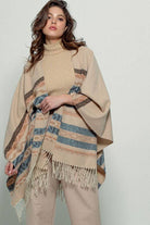 Woman wearing a beige poncho with a  Tan background and light blue, brown, light rust, and grey aztec pattern design  in stripes with tassel trim along bottom hem. Light weight fabric 