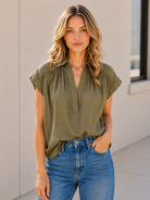 A woman wearing an muted olive green /khaki short-sleeve blouse with a gathered V-neckline, paired with blue jeans, standing against a light beige wall.