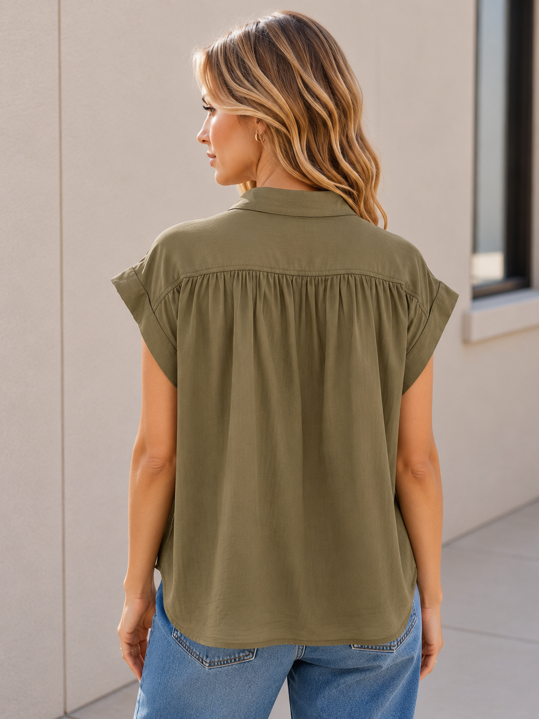 A woman shown from the back wearing an olive green /khaki short-sleeve blouse with a gathered back seam, paired with blue jeans, standing against a light beige wall.