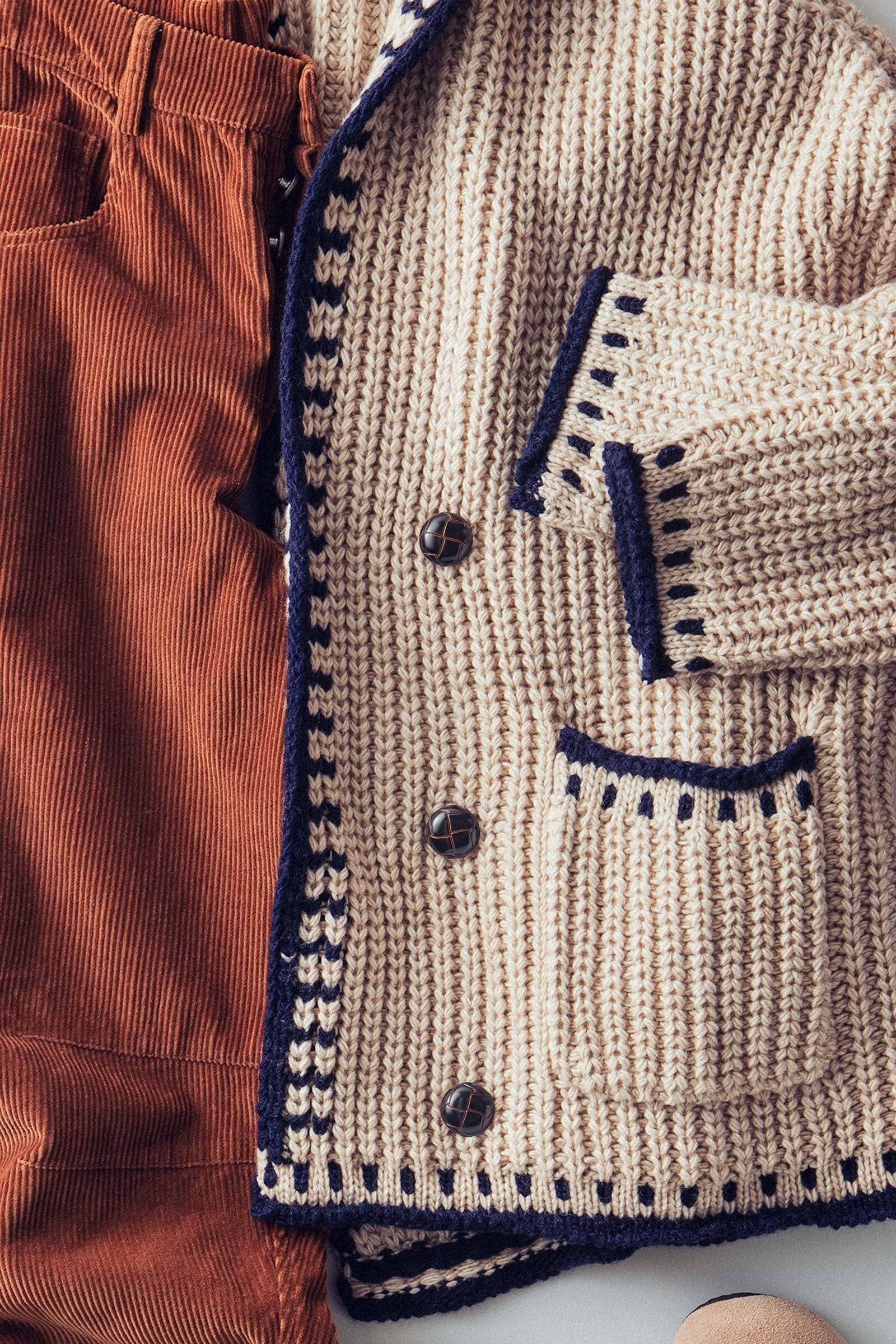 Close-up of a beige knitted cardigan with navy trim and buttons, paired with brown pants.