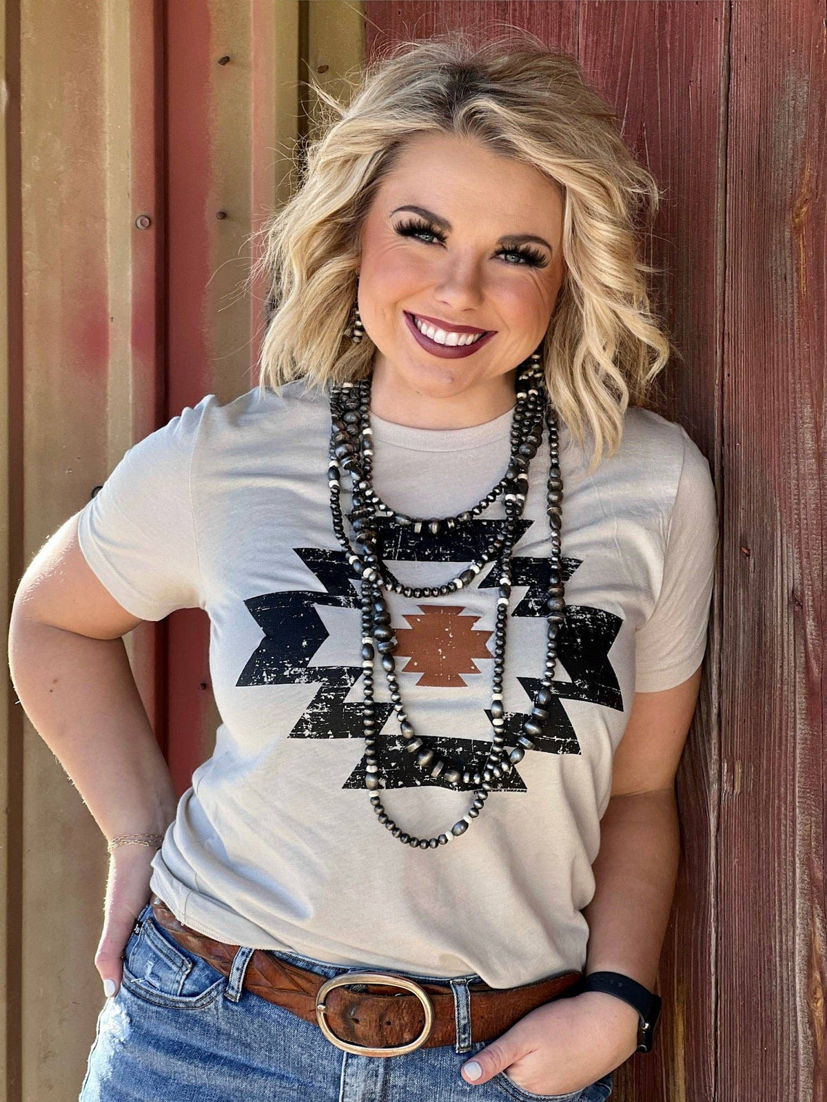 Woman wearing a white t-shirt with black geometric patterns, standing against a wooden wall.