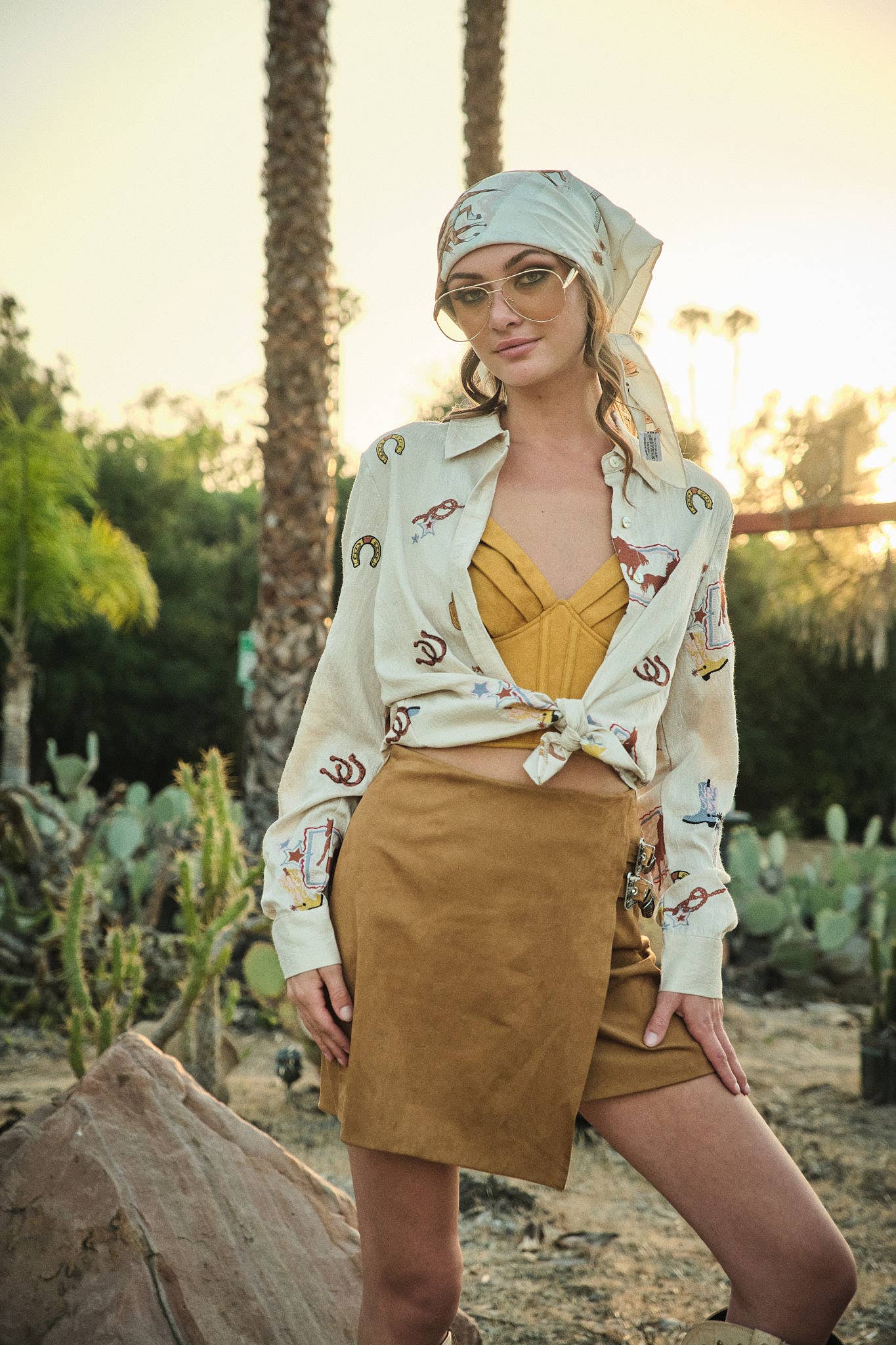 Woman in a desert setting wearing a patterned shirt, mustard top, and brown skirt.