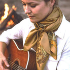 Woman playing guitar with a gold scarf around her neck, standing near a campfire.
