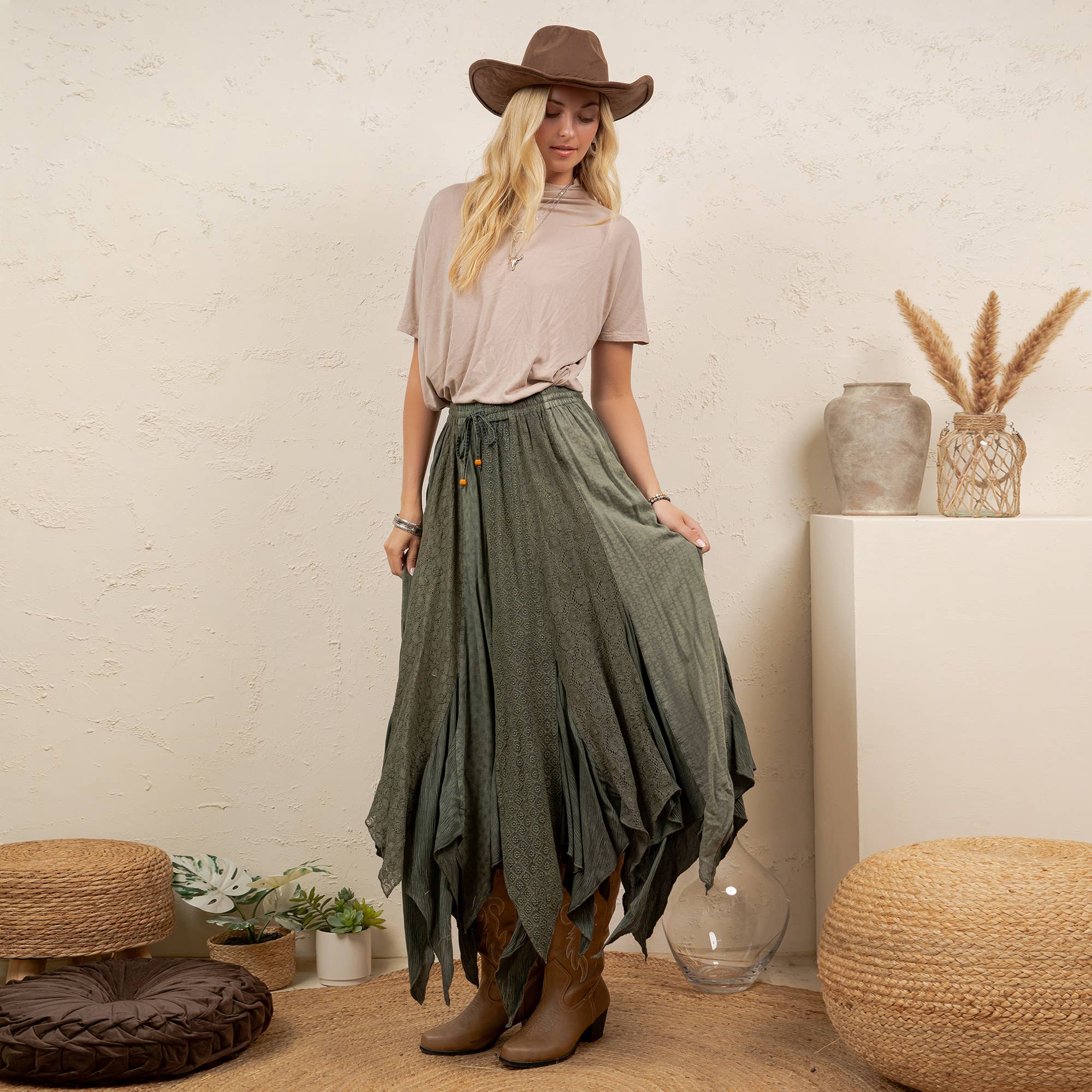 A woman stands in a styled indoor setting wearing a brown wide-brim hat, a light beige short-sleeve top, a long olive green tiered skirt, and brown ankle boots. The room has a textured cream wall, woven poufs, plants, and decorative vases.