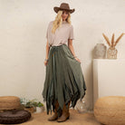 A woman stands in a styled indoor setting wearing a brown wide-brim hat, a light beige short-sleeve top, a long olive green tiered skirt, and brown ankle boots. The room has a textured cream wall, woven poufs, plants, and decorative vases.
