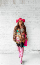 Person wearing a leopard print jacket, pink cowboy hat, and pink boots against a white brick wall.