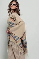 Woman wearing a beige poncho with a  Tan background and light blue, brown, light rust, and grey aztec pattern design  in stripes with tassel trim along bottom hem. Light weight fabric 