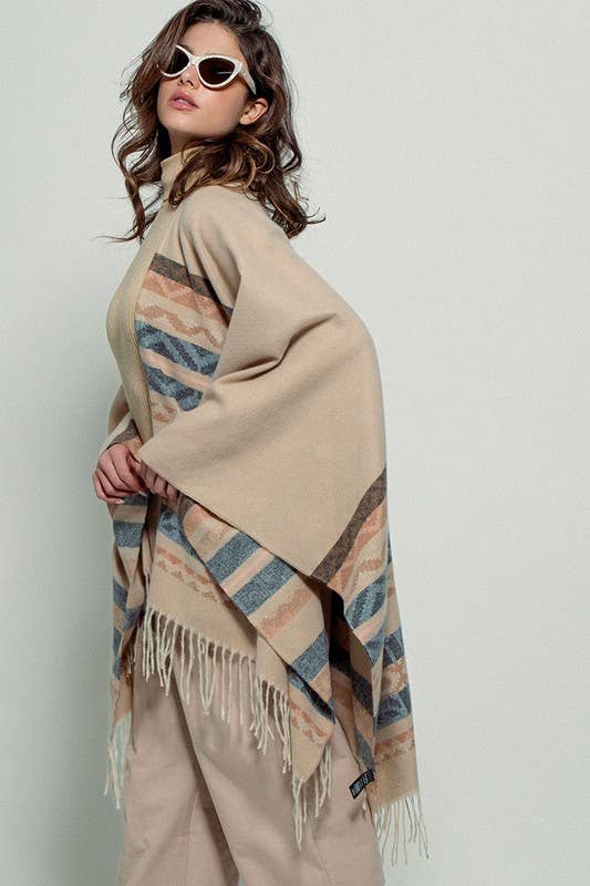 Woman wearing a beige poncho with a  Tan background and light blue, brown, light rust, and grey aztec pattern design  in stripes with tassel trim along bottom hem. Light weight fabric 
