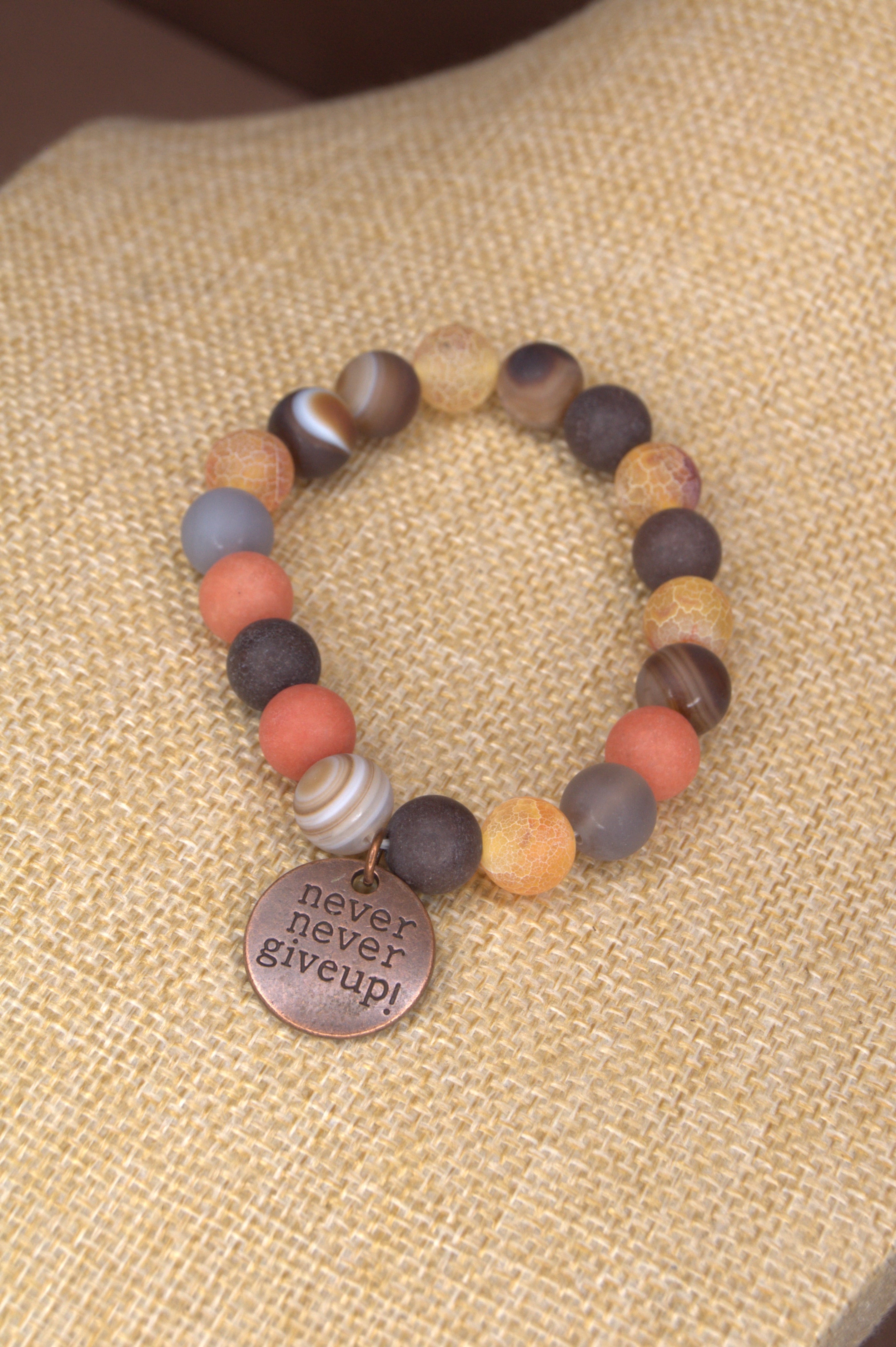 Beaded earth tone bracelet with a 'never never give up!' copper charm on a textured beige surface