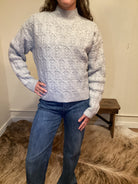 Person wearing a gray cable knit sweater and blue jeans standing against a neutral wall.
