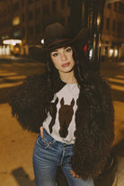 Woman wearing a cowboy hat, faux fur jacket, and horse-themed shirt at night.