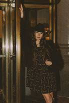 Woman in a patterned dress and fur coat standing in front of a mirror.