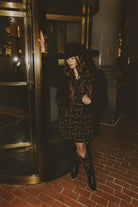 Person wearing a black coat, hat, and patterned dress standing in a dimly lit hallway.