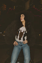 Woman wearing a white sweater with a horse design, brown faux fur jacket and blue jeans at night.
