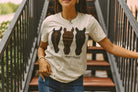 A woman wearing a cream-colored graphic t-shirt with three horse silhouettes in black and brown, standing on outdoor wooden stairs.