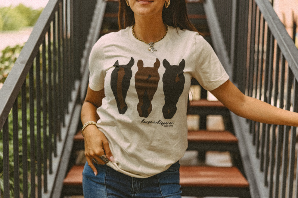 A woman wearing a cream-colored graphic t-shirt with three horse silhouettes in black and brown, standing on outdoor wooden stairs.