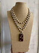 A multi-strand beaded necklace displayed on a burlap-covered mannequin bust. The necklace features cream-colored beads with small metallic spacer beads, and a dark reddish-brown rectangular pendant with white dot details.