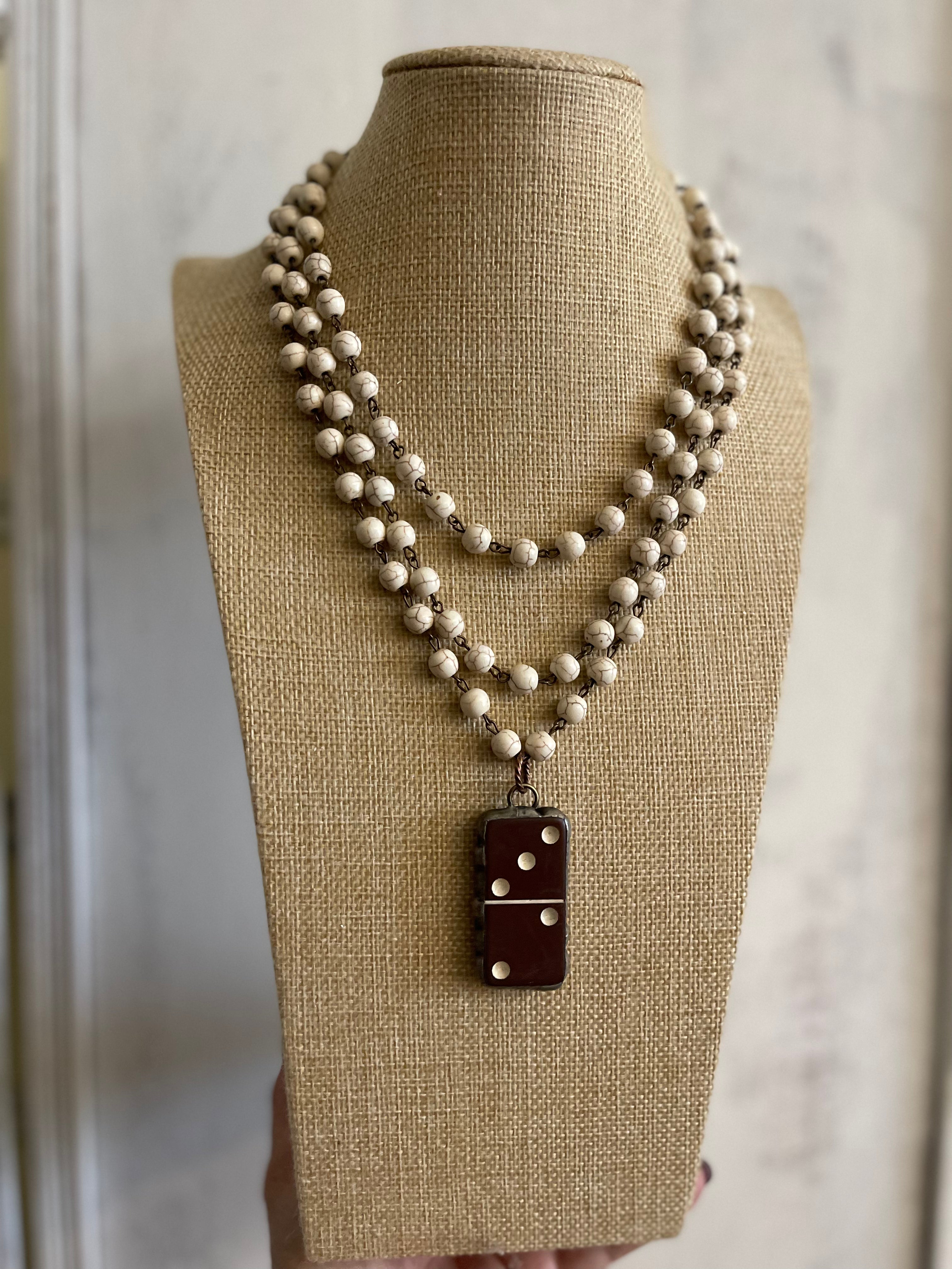 A multi-strand beaded necklace displayed on a burlap-covered mannequin bust. The necklace features cream-colored beads with small metallic spacer beads, and a dark reddish-brown rectangular pendant with white dot details.