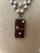 A close-up of a necklace with a dark brown domino-shaped pendant and white marbled beads on a chain, displayed on a beige burlap background.