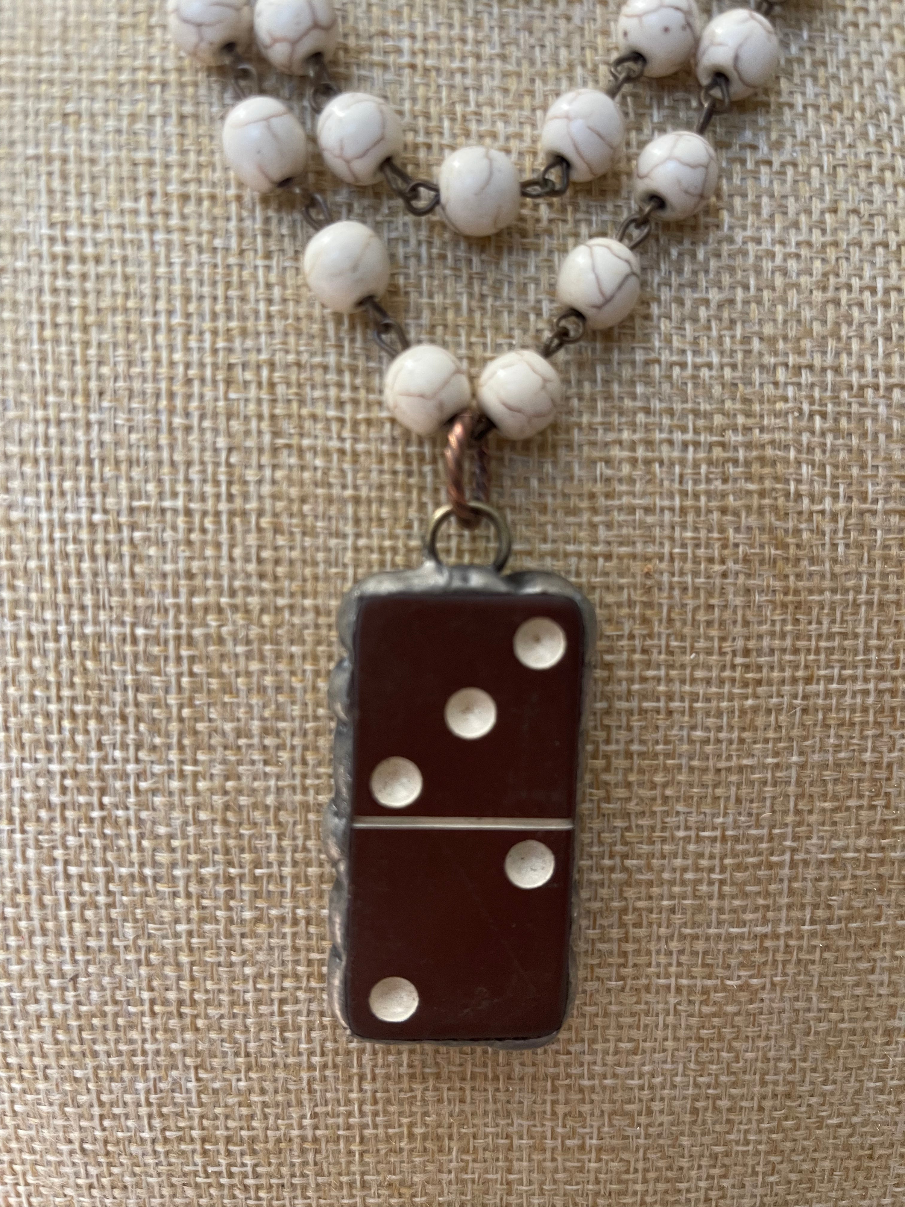 A close-up of a necklace with a dark brown domino-shaped pendant and white marbled beads on a textured beige fabric background.