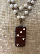 A close-up of a necklace with a dark brown domino-shaped pendant and white marbled beads on a textured beige fabric background.