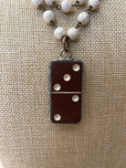 A close-up of a necklace with a dark brown domino-shaped pendant and white marbled beads on a chain, displayed on a beige burlap background.