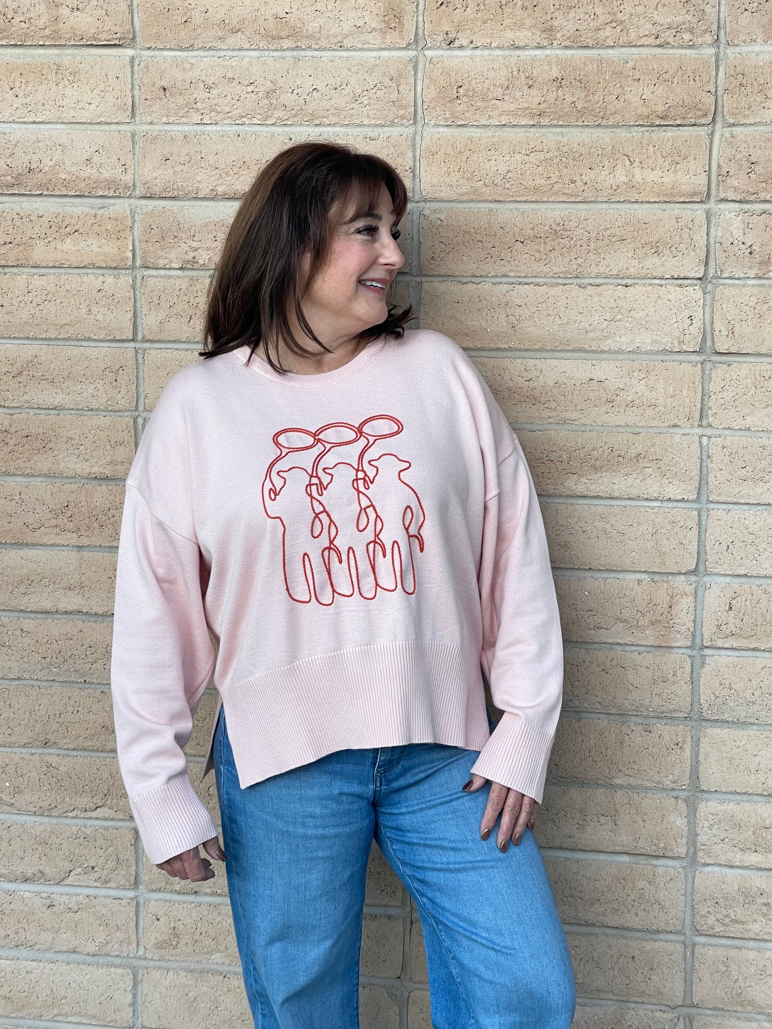 A person wearing a light pink crewneck sweatshirt with a orange line-art embroidery graphic of three cowboy figures, paired with blue jeans, standing against a light tan brick wall.