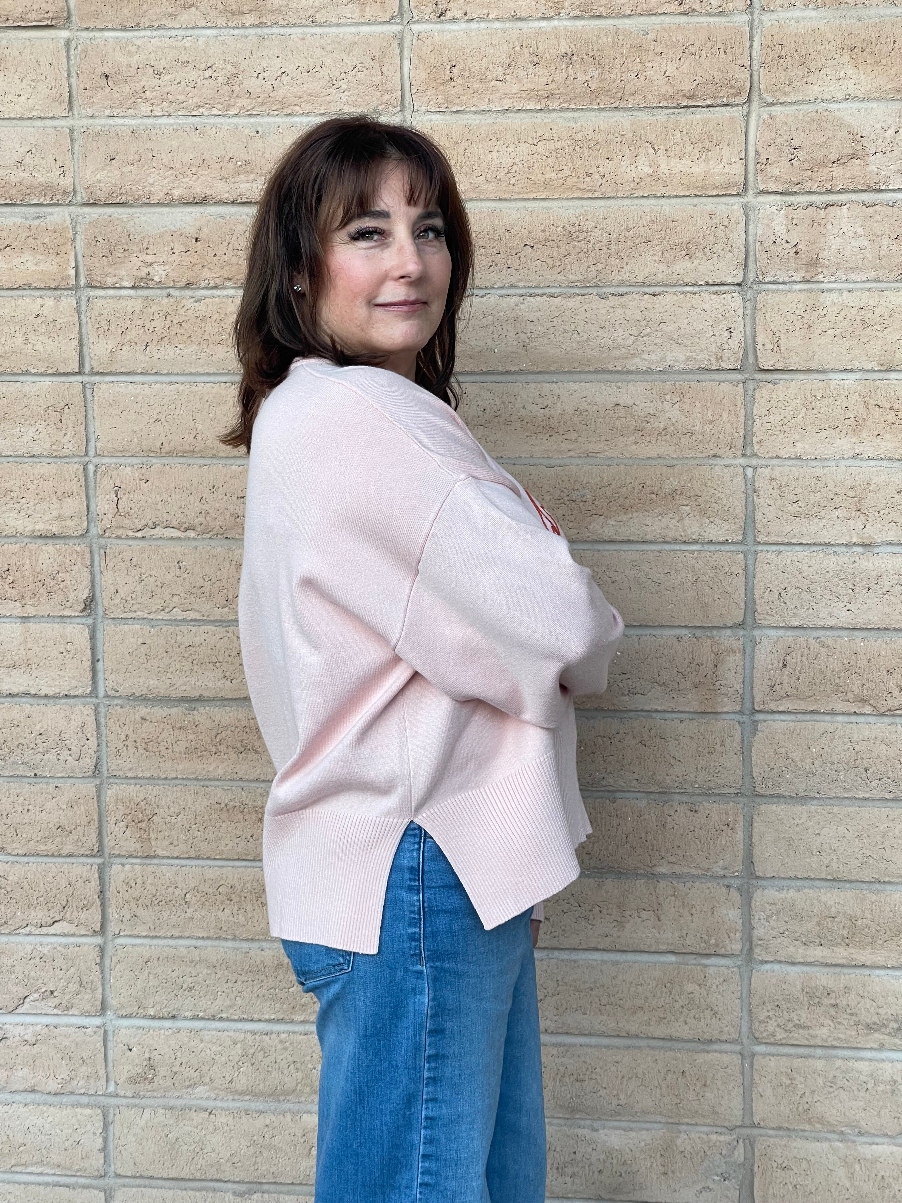 A woman with brown hair stands against a beige brick wall, wearing a light pink ribbed knit sweater and blue jeans.