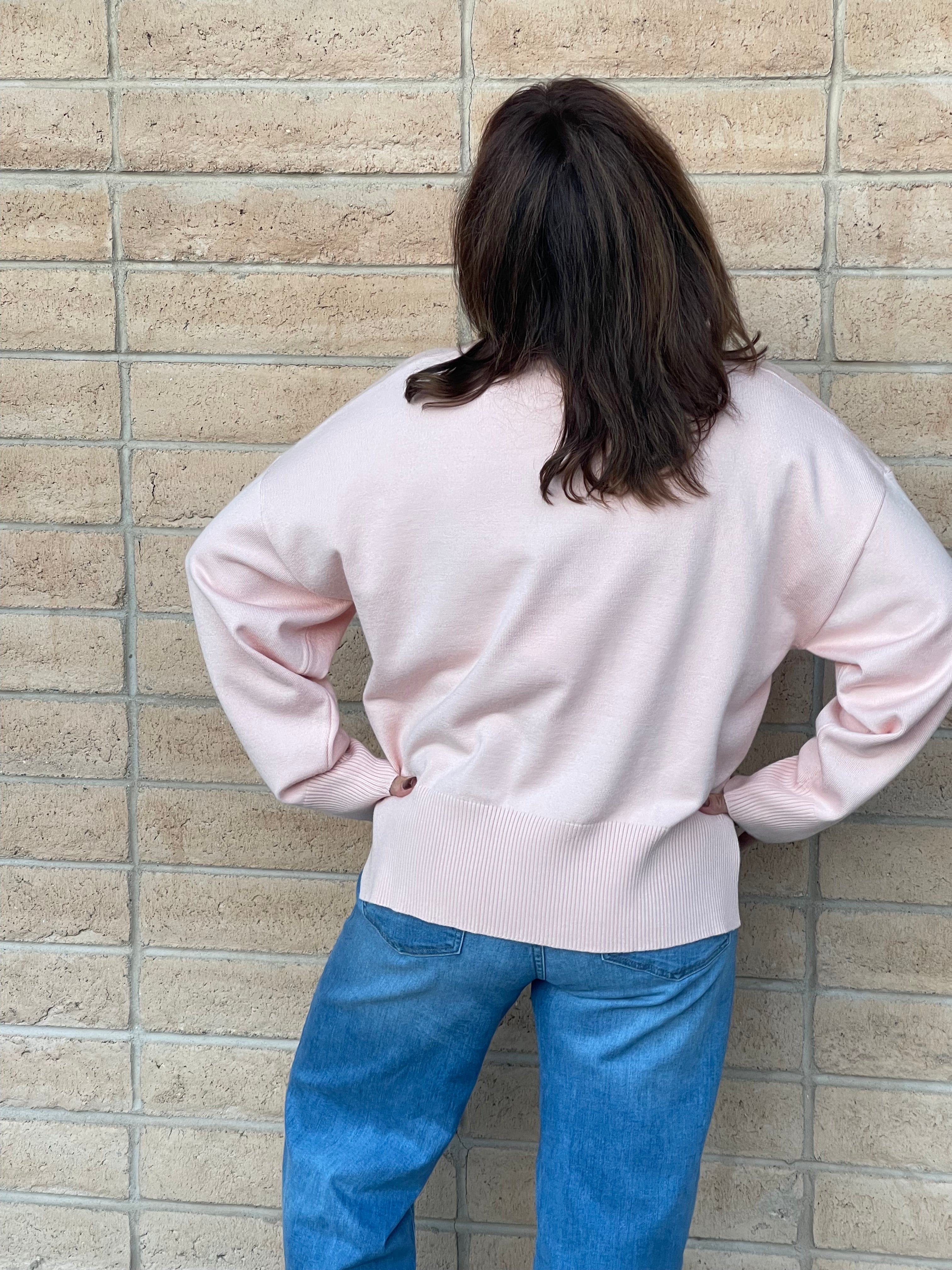 A person viewed from behind wearing a light pink knit sweater and blue jeans, standing against a beige brick wall.