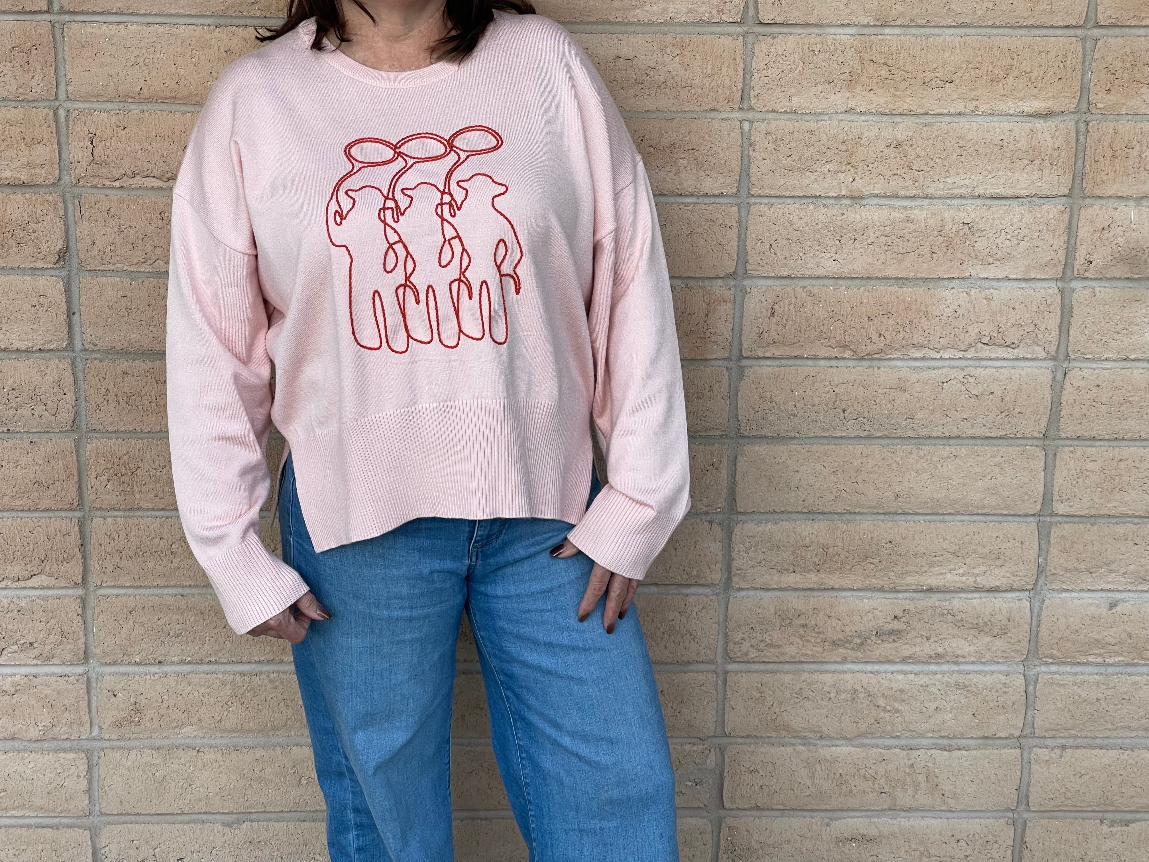 A person wearing a light pink crewneck sweatshirt with a orange line-art embroidery graphic of three cowboy figures, paired with blue jeans, standing against a light tan brick wall.