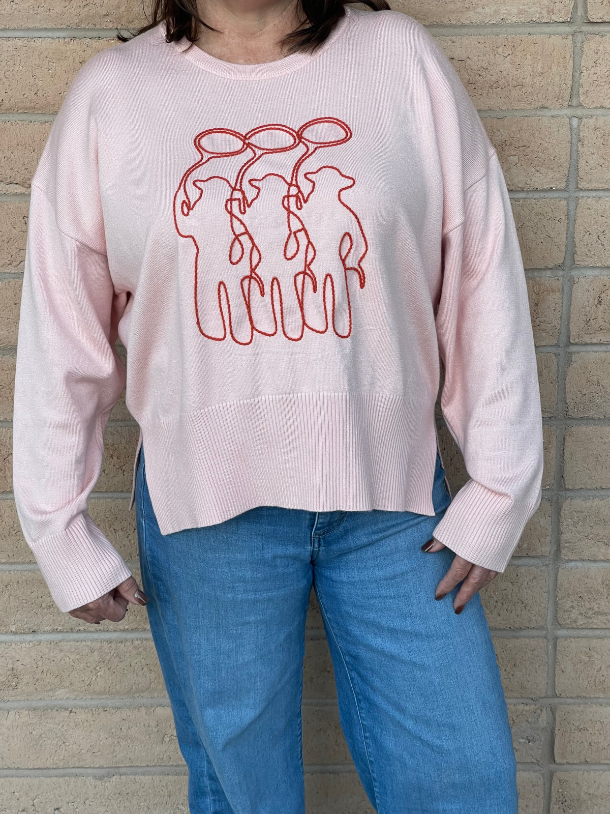 A person wearing a light pink crewneck sweatshirt with a orange line-art embroidery graphic of three cowboy figures, paired with blue jeans, standing against a light tan brick wall.