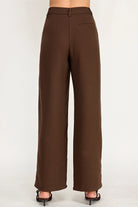 A person shown from the back wearing high-waisted, wide-leg brown trousers and black heeled sandals against a plain light gray studio background.