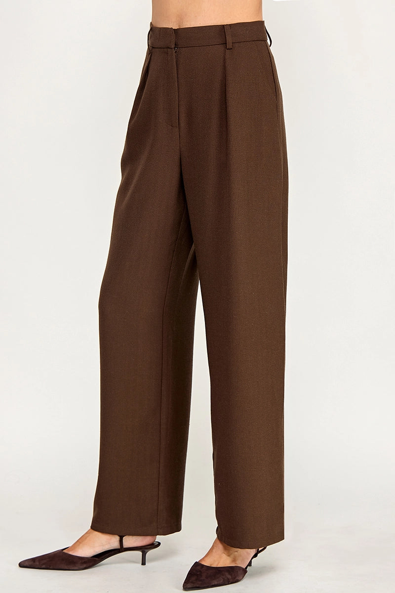 A person wearing high-waisted, wide-leg brown trousers with a matching belt, styled with dark brown pointed-toe slingback heels against a plain light gray studio background.