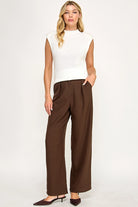 A woman modeling a white sleeveless mock-neck knit top and dark brown wide-leg trousers, paired with dark brown pointed-toe heels, against a plain white studio background.