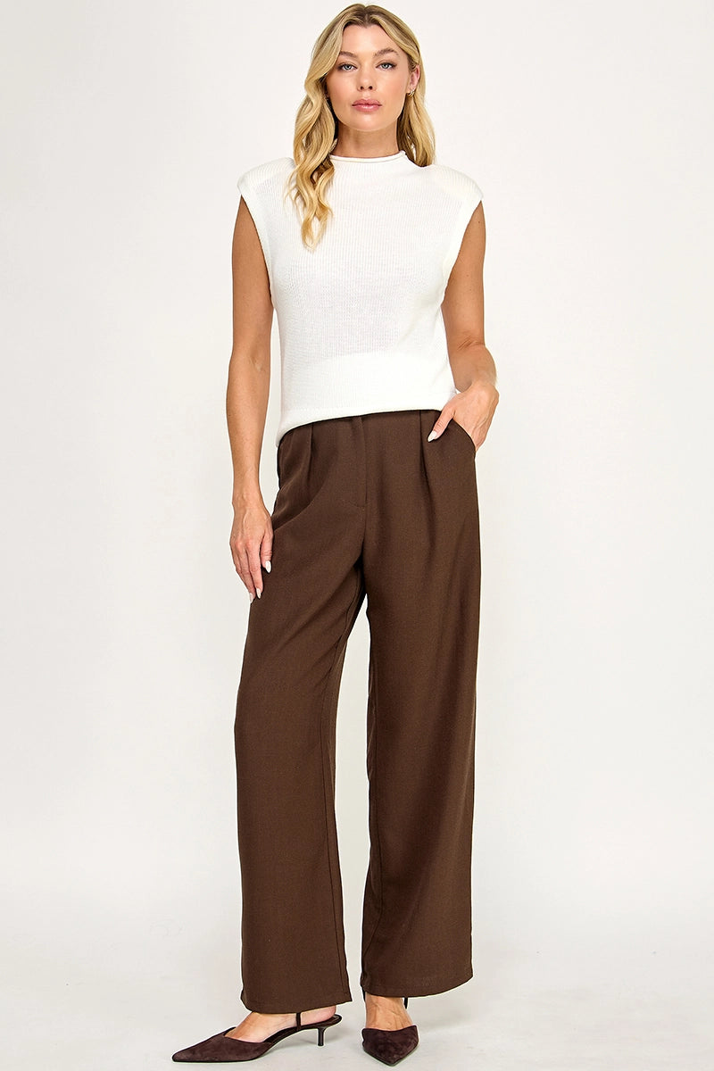 A woman modeling a white sleeveless mock-neck knit top and dark brown wide-leg trousers, paired with dark brown pointed-toe heels, against a plain white studio background.