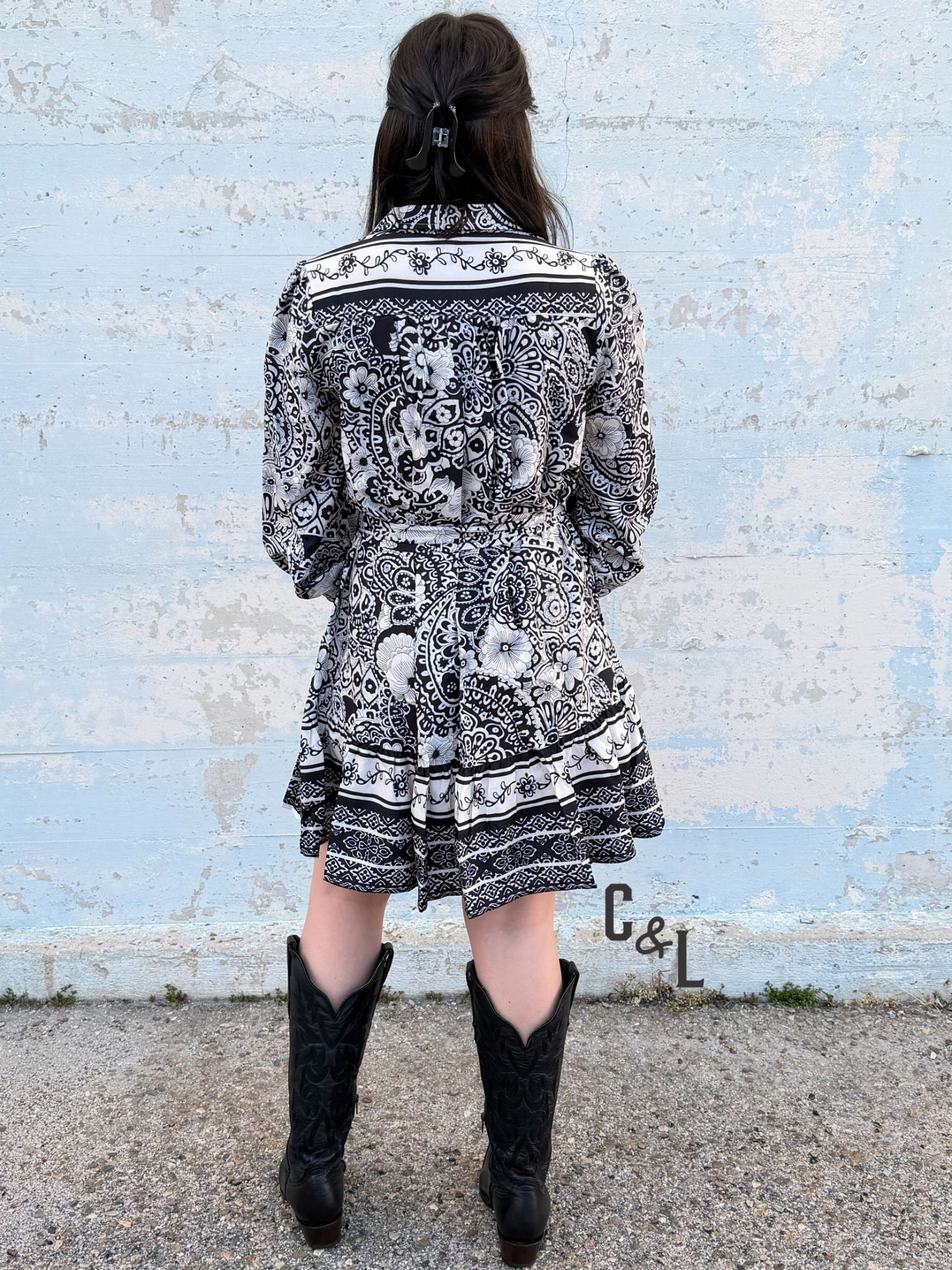 Person wearing a black and white patterned dress with cowboy boots standing against a light blue brick wall.