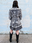 Person wearing a black and white patterned dress with cowboy boots standing against a light blue brick wall.