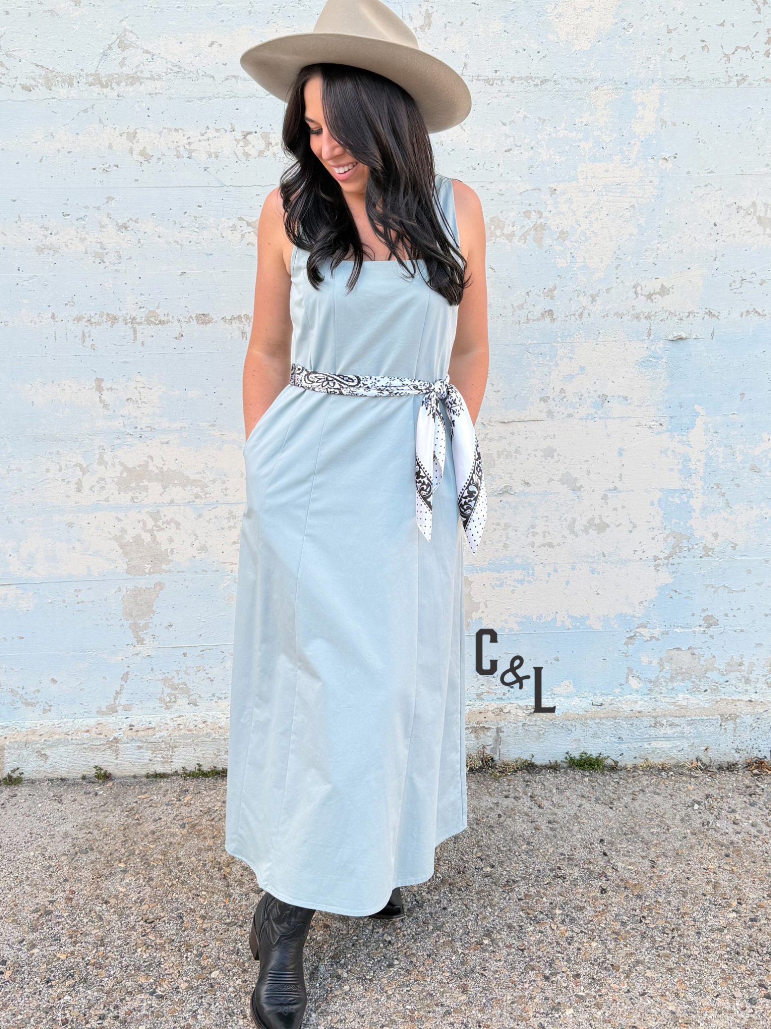 Woman wearing a light blue dress with a belt and a hat, standing against a textured wall.