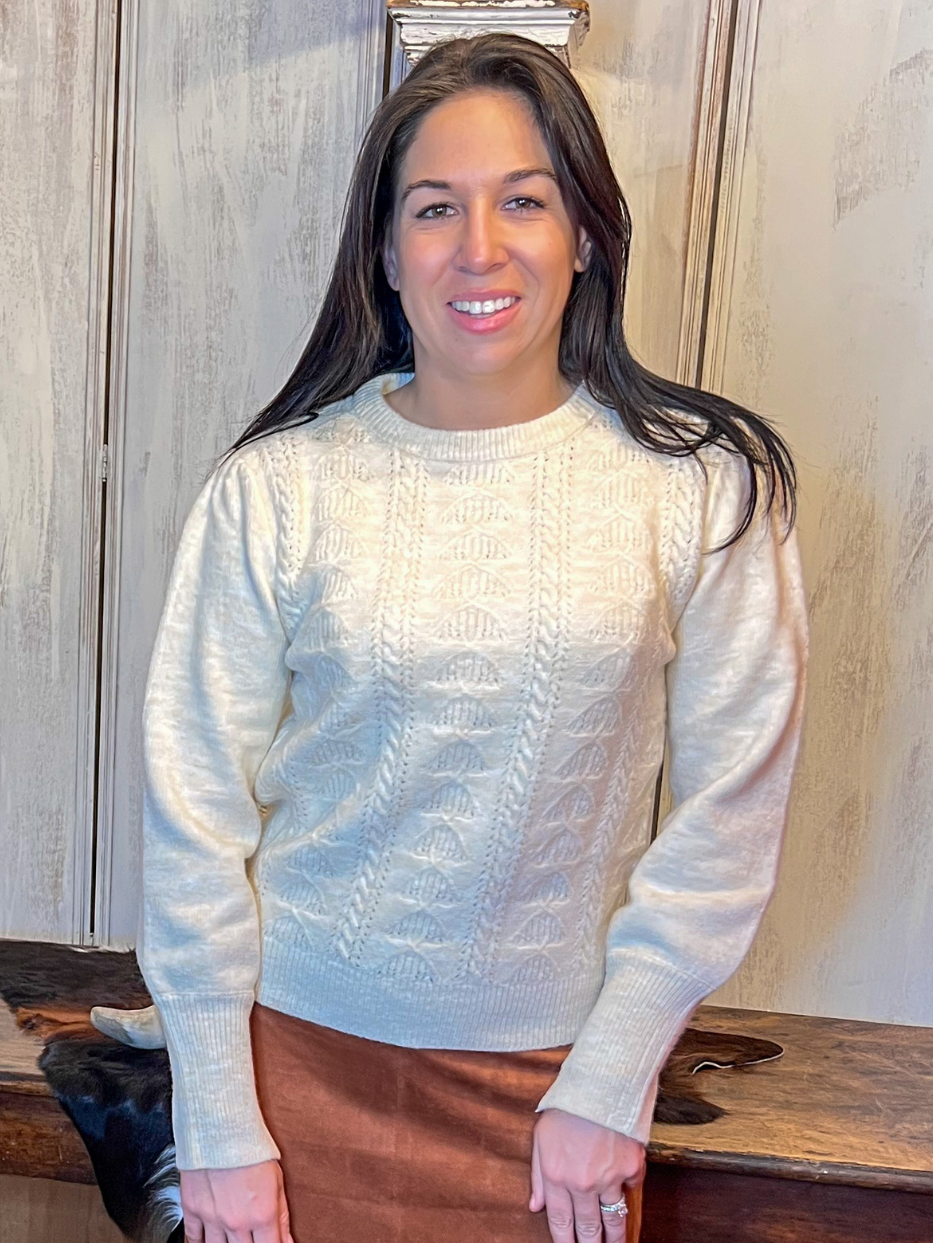 Woman wearing a light sweater and brown skirt standing in a room with wooden furniture.