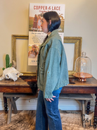 Person wearing a green jacket and blue jeans standing in front of a table with decorative items and posters.