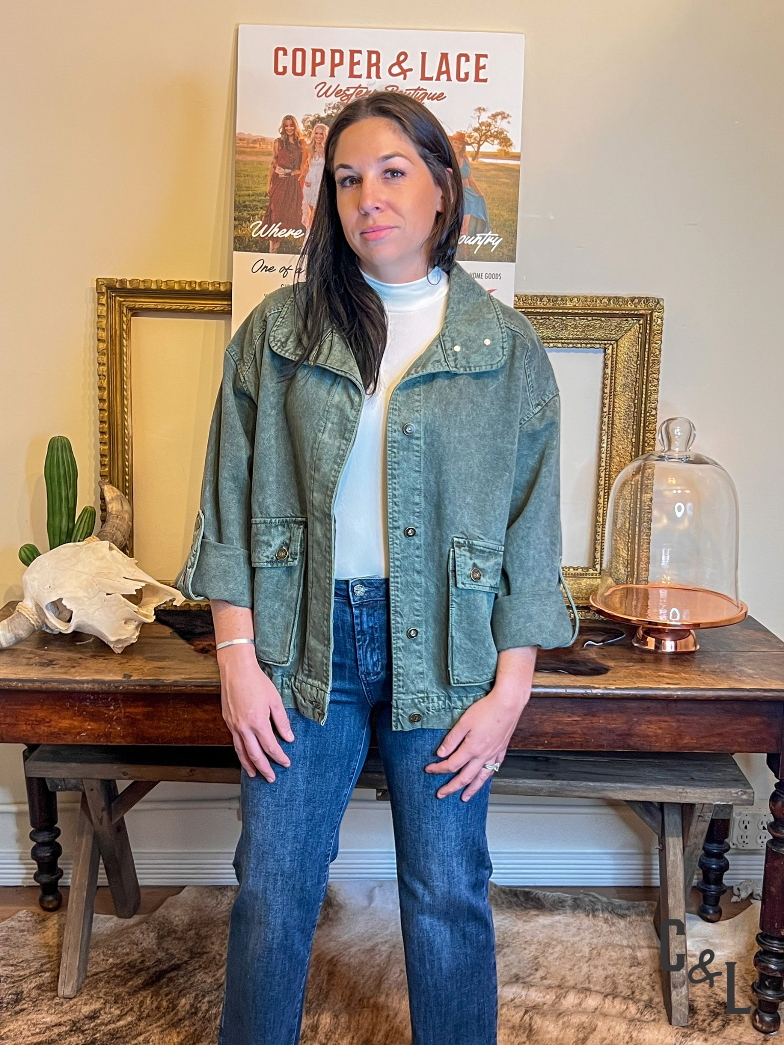 Woman wearing a green jacket and blue jeans standing in front of a table with decorative items.