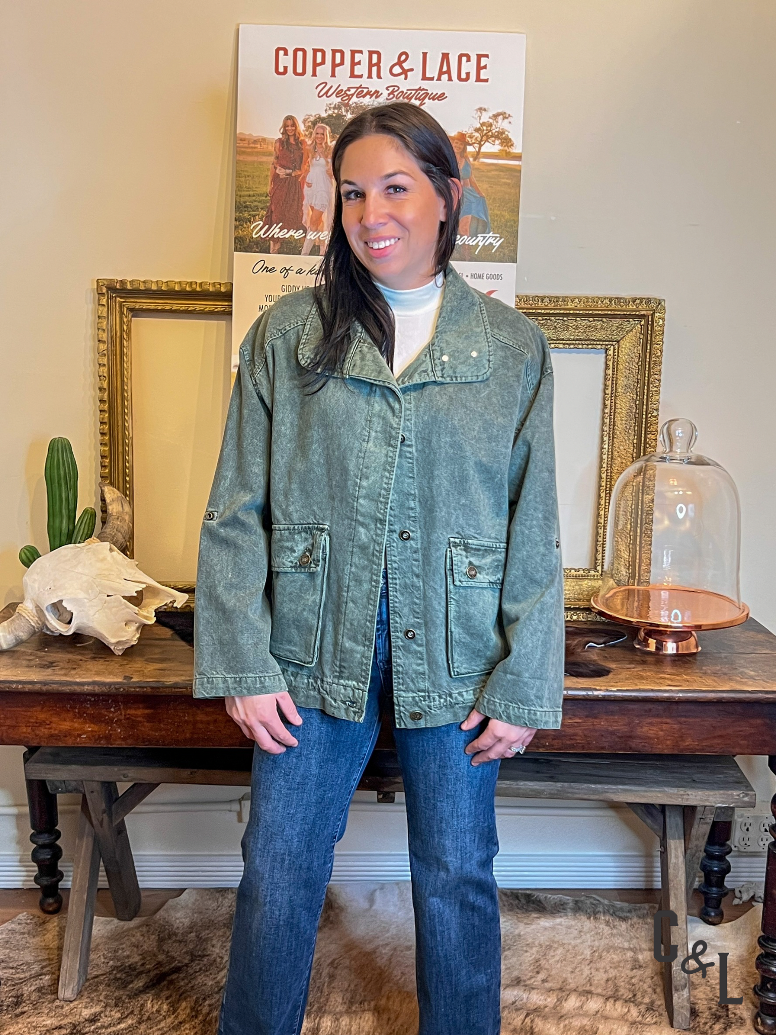 Woman wearing a green jacket denim bomber jacket and blue jeans standing in front of a 'Copper & Lace' sign.