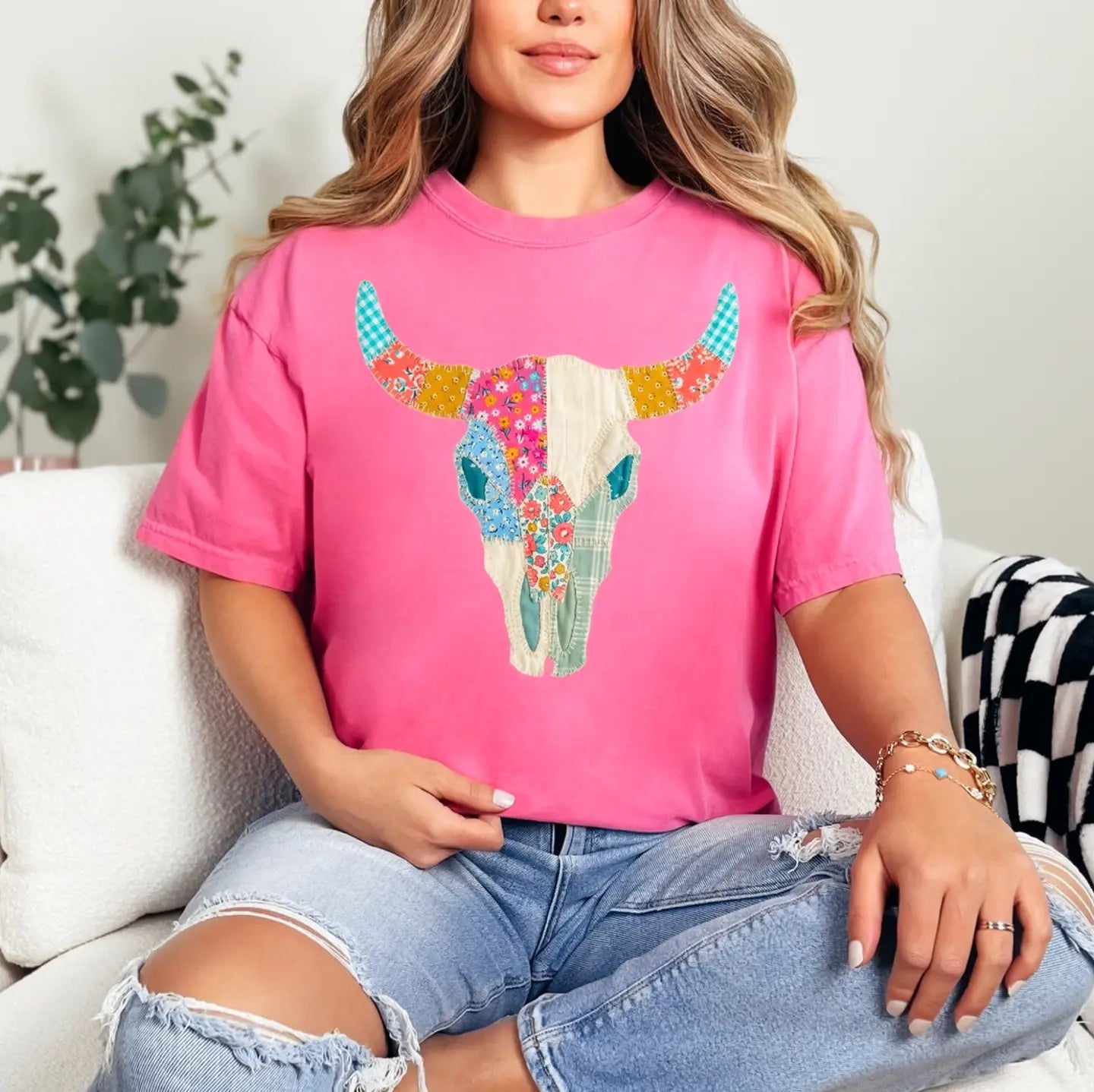 A woman wearing a bright pink t-shirt with a colorful patchwork bull skull graphic, paired with distressed light blue jeans, sitting on a white couch.