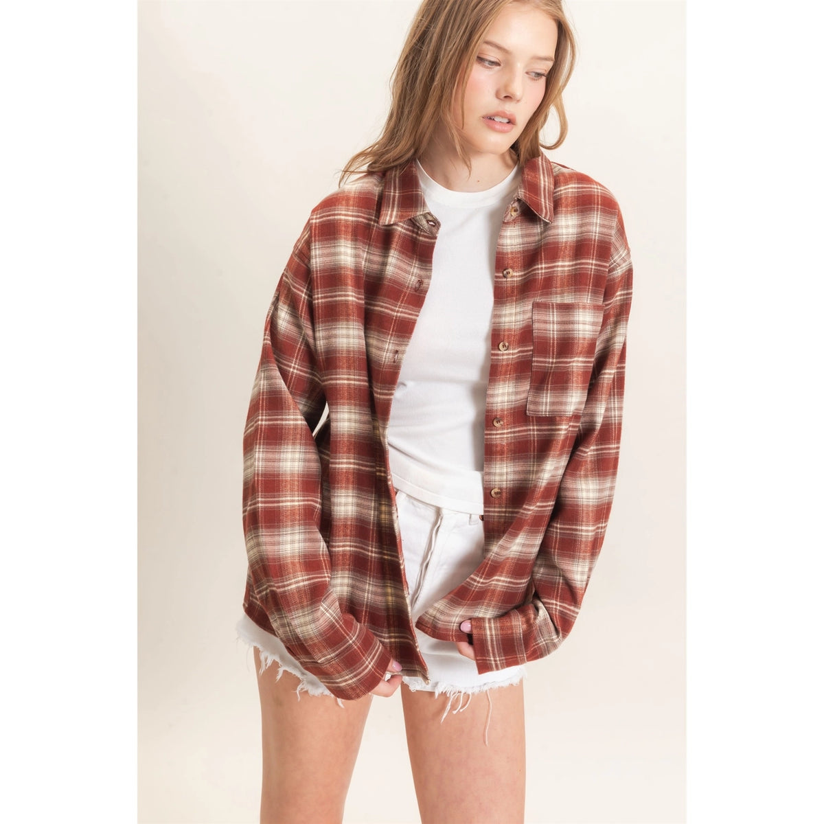 A woman wearing an oversized rust red and beige plaid flannel shirt over a white t-shirt and white frayed denim shorts, standing against a plain off-white background.