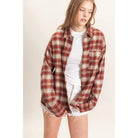 A woman wearing an oversized rust red and beige plaid flannel shirt over a white t-shirt and white frayed denim shorts, standing against a plain off-white background.